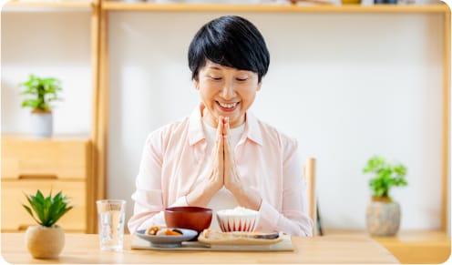 テーブルに座り、手を合わせて感謝の祈りを捧げる女性。食卓には和食が並び、背景には植物が飾られている。